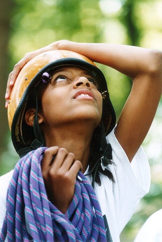 The image shows a young person wearing a helmet and holding a rope. They are looking upwards with a thoughtful expression. The person appears to be outdoors, possibly in a wooded area, as suggested by the blurred background. The lighting indicates it might be daytime.